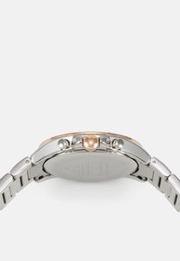 Reloj de acero inoxidable plateado con una caja redonda, acentos en oro rosado y una corona texturizada; presenta una pulsera de metal y un fondo liso.