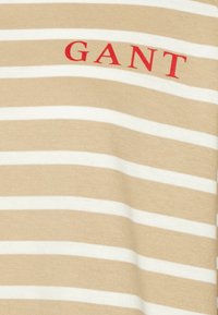 Gestreiftes T-Shirt in Beige und Weiß mit einem auffälligen roten "GANT"-Logo. Weicher, strukturierter Stoff mit horizontalen Streifen.