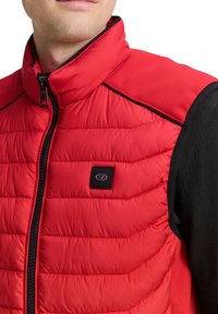 Rote Steppweste mit gestepptem Design und schwarzen Akzenten, mit hohem Kragen und Reißverschluss, präsentiert am Oberkörper eines Models.