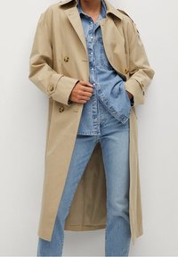 Trench-coat beige à double boutonnage, grands boutons et col classique, superposé à une chemise en denim bleu clair et un jean.