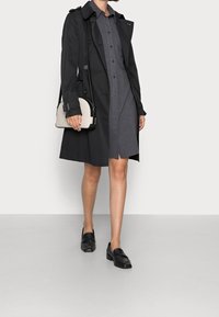 Czarny płaszcz trench nałożony na szary sukienkę, w połączeniu z czarnymi mokasynami i trzymaniem jasnoszarej torby na ramię z czarnymi akcentami.