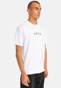 White cotton t-shirt with short sleeves and a crew neckline, featuring a graphic design on the chest in black. Worn with dark jeans.