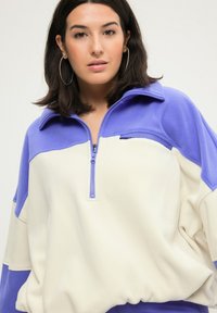 Fleece-Pullover mit halbem Reißverschluss, in Lavendel- und Cremefarbblöcken, mit Stehkragen und lässiger Passform. Weicher Stoff und legere Gestaltung.