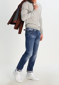 Graues Langarmshirt, blaue Jeans, weiße Sneakers. Braune Jacke über der Schulter. Die Jeans haben einen leichten Fade-Effekt und aufgerollte Säume.