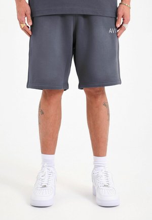 Hombre con pantalones cortos gris oscuro con el logo "AVN", zapatillas blancas, calcetines blancos y tatuajes visibles en las piernas, de pie sobre un fondo blanco.