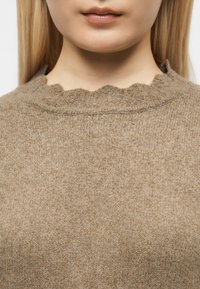 Gros plan sur le bas du visage et le cou d'une personne portant un pull en maille marron clair avec un col festonné.