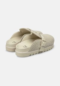 Scarpe slip-on beige intrecciate con una tomaia strutturata e una larga fascia con fibbia metallica, poggiate su una suola spessa e dentata.