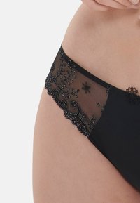 Czarne majtki z przezroczystymi koronkowymi bocznymi panelami, zdobionymi misternym haftem kwiatowym i koralikami, kontrastującymi z gładką czarną tkaniną.