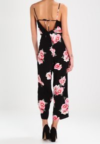 Combinaison noire à motifs floraux roses, bretelles spaghetti et dos nu. Composée de jambes larges et d'un corsage ajusté. Ornée d'un nœud papillon.