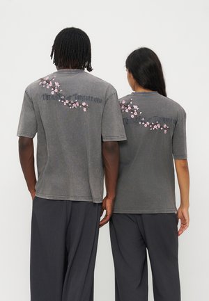 Deux personnes portant des t-shirts gris avec une broderie florale rose et le texte "Threads of Tomorrow" dans le dos, debout côte à côte, dos à nous.