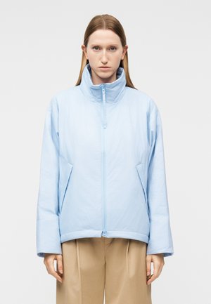 Femme aux cheveux lisses châtain clair porte une veste matelassée light blue à fermeture éclair et un pantalon beige à jambes larges, debout devant un fond clair uni.