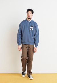 Pull-over bleu marine avec un col et un logo sur la poitrine, associé à un pantalon marron et des baskets noires. Texture lisse, design décontracté.