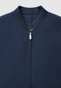 Veste bomber bleu marine avec un col côtelé et une fermeture éclair argentée au centre devant, sur un fond clair.