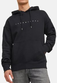 Sort hættetrøje med kængurulomme, prydet med hvid tekst "JACK & JONES" centreret på brystet. Snore med metalspidser og ribbede manchetter.
