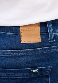 Jeans din denim cu o etichetă din piele maro embosată cu "MUSTANG" și un mic logo cu un cal. Jeansii sunt de o nuanță albastră închisă, cu o textură subtilă.