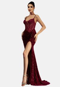 Frau in einem burgunderfarbenen Paillettenkleid mit hohem Schlitz und Spitzen-Corsage, die mit den Händen in der Hüfte posiert und silberne High-Heels trägt.