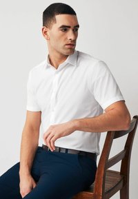 Camisa blanca de manga corta con cuello estándar y botones, combinada con pantalones azul marino. Textura de tela suave, diseño minimalista.