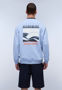 Lys blå hettegenser med et grafisk design på baksiden, med svart-hvite bølgemønstre og teksten "NAPAPURI" over og "geographic" under.