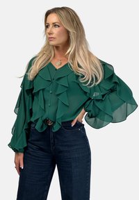 Szmaragdowo zielona bluzka z falbanami, z długimi rękawami i zapinana na guziki, w zestawie z ciemnoniebieskimi dżinsami i złotym paskiem akcentującym.