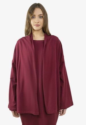 Młoda kobieta ubrana w luźną, długorękawową bordową kurtkę nałożoną na dopasowaną sukienkę, stojąca na jednolitym tle.