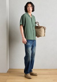 Green short-sleeve shirt with vertical stripes, blue denim jeans, beige slip-on shoes, and a tan bag with brown accents. Wood floor backdrop.