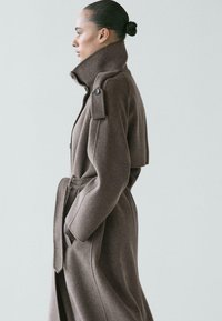 Donna di profilo che indossa un lungo cappotto marrone di lana con cintura, colletto alto e spalline con bottoni, capelli raccolti in uno chignon stretto su sfondo semplice.