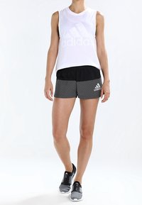 Vit ärmlös tanktopp med subtil logotyp, svarta träningsshorts med diagonala ränder och grå sneakers med dragkedjedetalj.