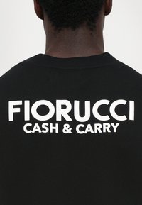 Zadný pohľad na osobu v čiernom tričku s veľkým bielym nápisom "FIORUCCI CASH & CARRY" potlačeným na hornej časti chrbta.