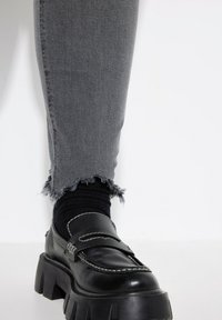 Mocasines de cuero negro con suelas gruesas, costuras blancas, combinados con jeans grises desgastados y calcetines negros, mostrando un look casual en capas.