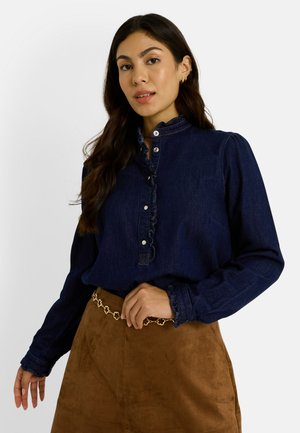Chemise en denim bleu foncé avec un col volanté, des manches longues et des boutons, associée à une jupe en suède marron et une ceinture décorative en or.