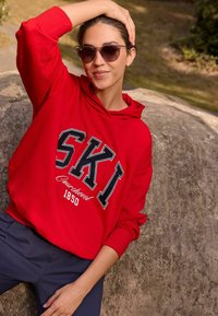 Sudadera roja con el logo "SKI" en letras negras en negrita, bolsillo tipo canguro y puños de canalé, combinada con pantalones oscuros. Con un fondo rocoso.