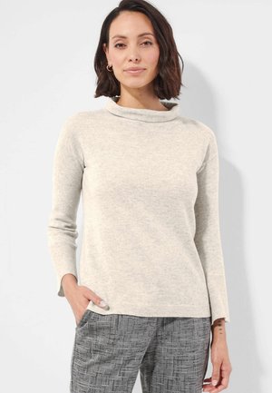 Femme aux cheveux courts et foncés portant un pull beige clair à manches longues et un pantalon gris à motifs, debout avec une main dans la poche.