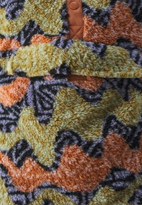 Fleecová oděv s vlnitým vzorem v oranžové, zelené a černé barvě. Obsahuje hnědý patentový knoflík u výstřihu. Měkká, texturovaná látka.