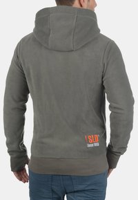Gray fleece hoodie with a high collar, visible stitching, and an orange logo on the lower back, featuring a standard fit and long sleeves.