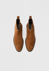 Braune Wildleder-Ankelboots mit glatter Textur, spitzem Zeh, schwarzen Sohlen und einem seitlichen Reißverschluss für einfachen Zugang. Stabiler Absatz mit Zuglasche.