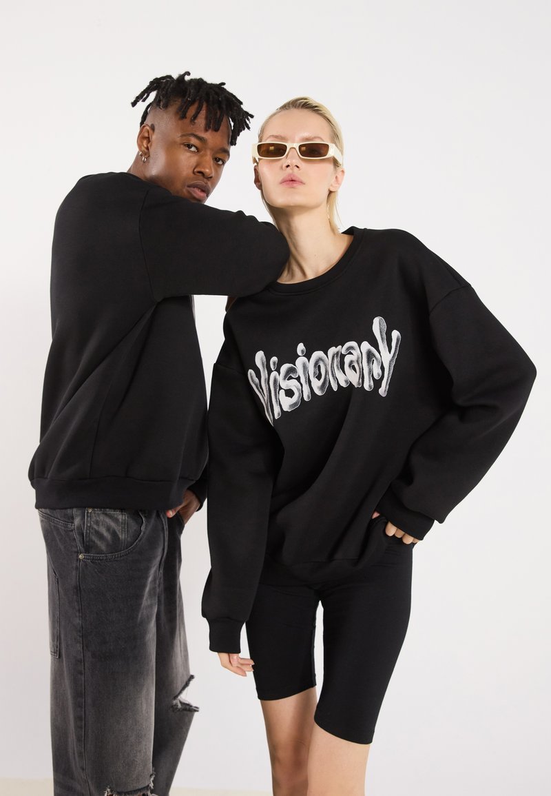 Due modelli posano in abbigliamento nero. Uno indossa una felpa nera oversize con la scritta "Visionary" in bianco, l'altro ha una felpa nera abbinata e dei pantaloni corti.