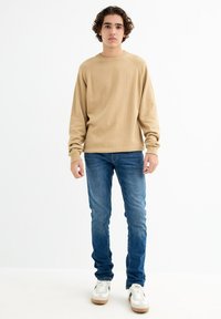 Suéter de manga larga color beige claro con un cuello acanalado, combinado con jeans ajustados azules y zapatillas blancas con acentos grises.