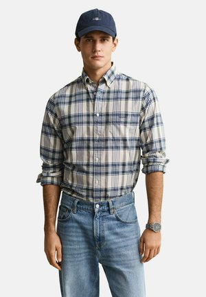 Mann trägt marineblaue Mütze, blau-beiges kariertes Hemd mit hochgekrempelten Ärmeln, hellblaue Jeans und eine silberne Armbanduhr vor weißem Hintergrund.