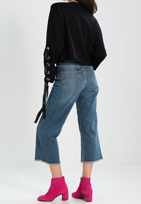Mujer vestida con una blusa negra de manga larga con cordones en las mangas, jeans cortos azules y botas de tobillo rosa brillante de pie contra un fondo sencillo.