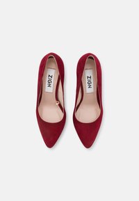 Escarpins à bout pointu en daim rouge avec une texture lisse et un design échancré. L'intérieur est doté d'une doublure claire, et la hauteur des talons est modérée.