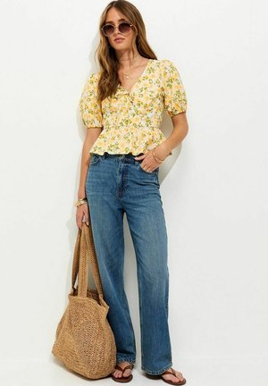 Vrouw met een gele bloemenpofmouw top, blauwe jeans, zonnebril en sandalen, die een grote geweven draagtas vasthoudt, staand tegen een witte muur.