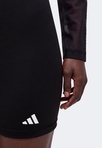 Personne portant un short Adidas noir avec logo blanc, debout le bras le long du corps et la main détendue près de la cuisse, sur un fond uni clair.