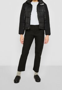 Chaqueta de abrigo negra con cremallera y capucha, camiseta blanca debajo, pantalones negros y zapatos oscuros con detalles texturizados.