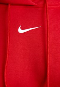 Piros Nike kapucnis pulóver fehér logóval. Puha anyagból készült, húzózsinórral ellátott kapucnival és varrási részletekkel rendelkezik. Simább textúra, minimális dizájn.