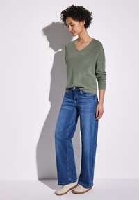 Maglione a V in maglia verde chiaro e luccicante; jeans blu a gamba larga; scarpe casual beige con suola testurizzata; modello in piedi contro uno sfondo neutro.