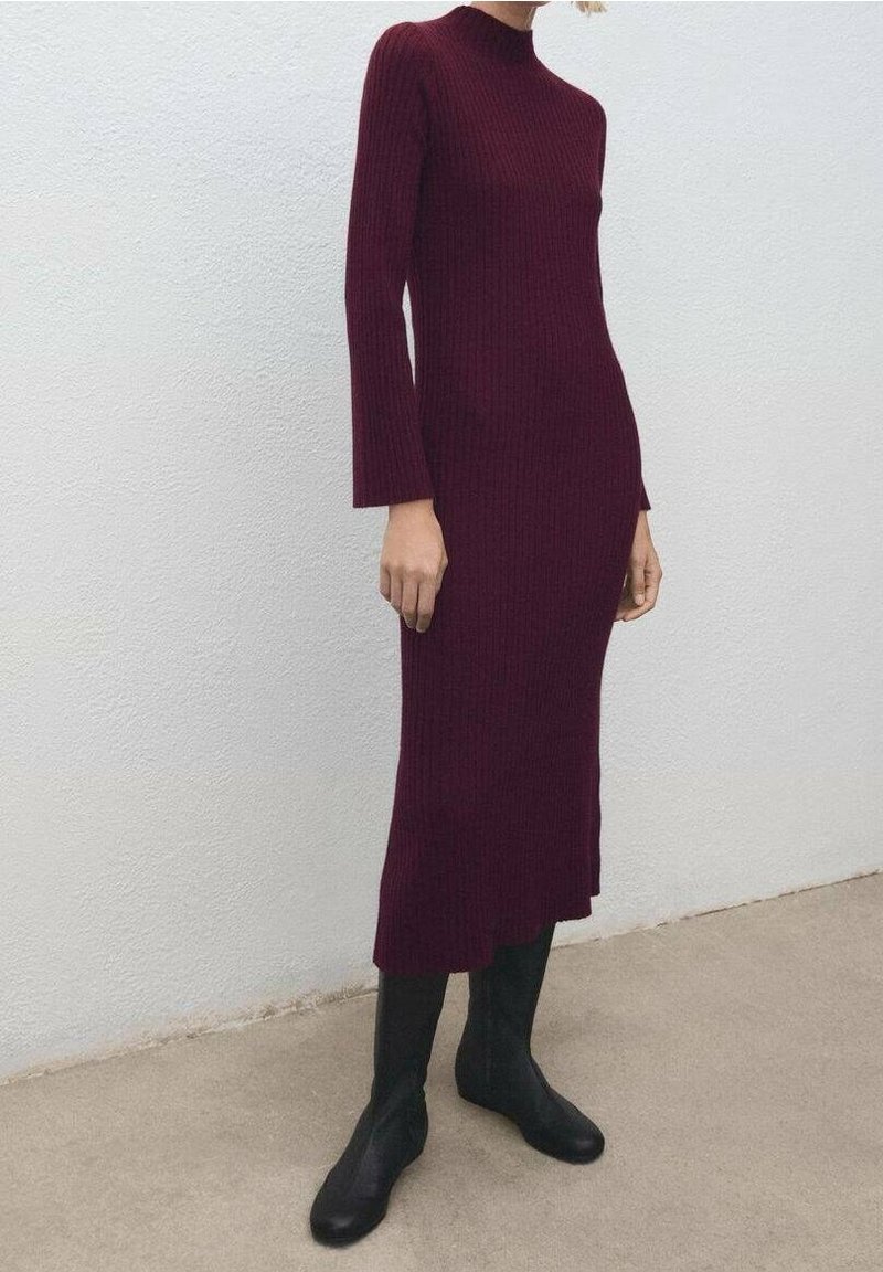 Femme portant une robe midi bordeaux à manches longues et côtelée, associée à des bottines noires, debout devant un mur blanc uni.
