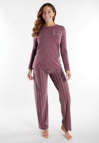 Woman smiling, wearing purple long-sleeve top with text and matching striped pants, standing barefoot on a white background.