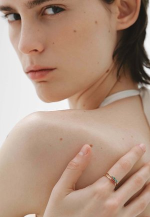 Anillos de oro con acentos verdes y rojos en una mano apoyada sobre un hombro, con piel suave y ligeras pecas.