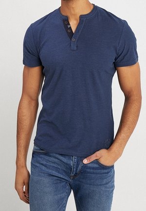 Uomo che indossa una maglietta henley blu navy aderente a maniche corte con patta con bottoni e jeans blu, mano nella tasca.