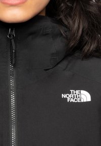 Giacca nera con zip completa, collo alto e logo bianco sulla parte sinistra del petto. Il materiale sembra liscio con una finitura opaca.
